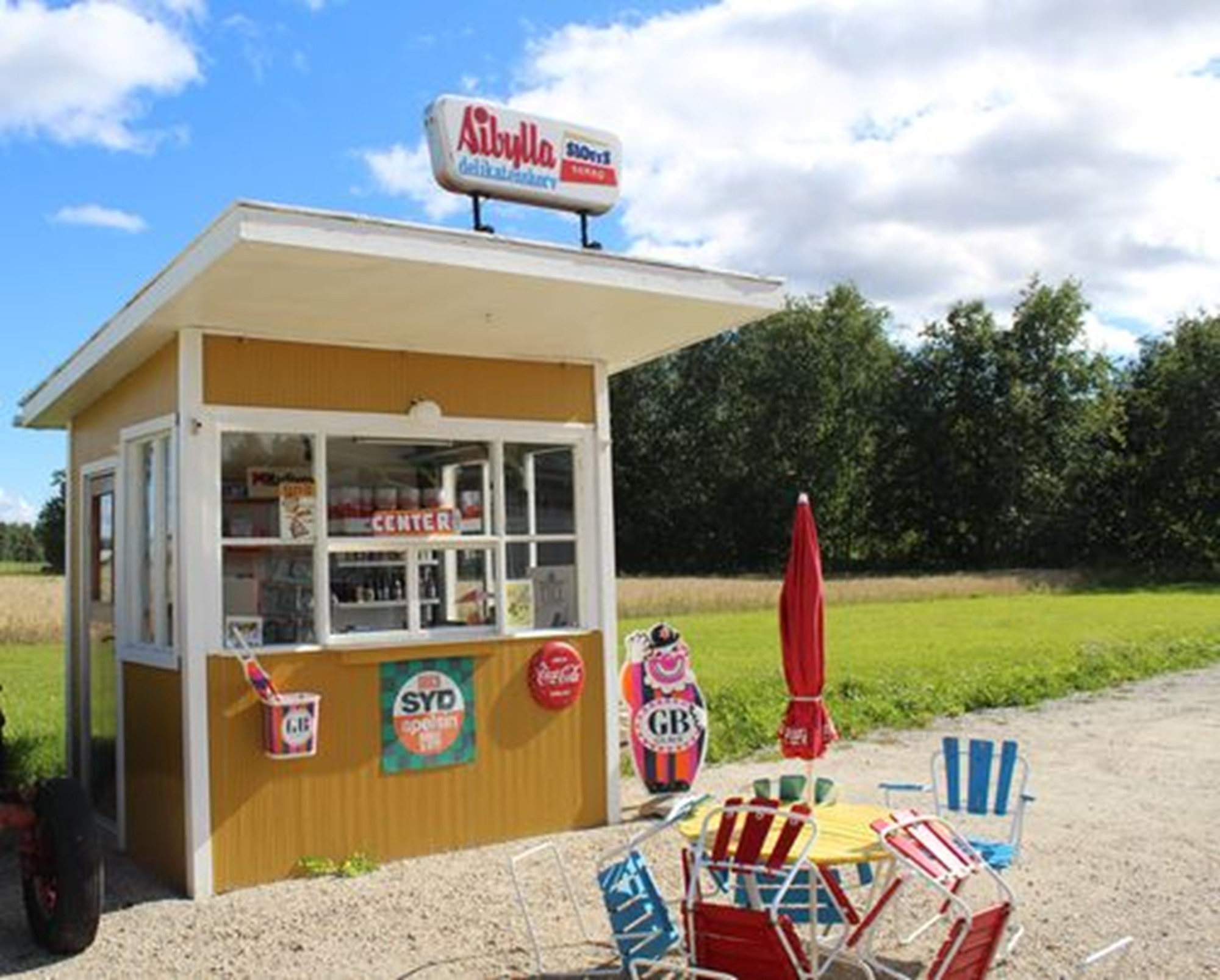 Här snackar vi Sommarlovs barndom Kiosken med alla de rätta sakerna, utemöblerna, blå himmel och gröna gräsmattor.